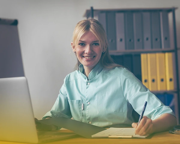 Женщина-врач сидит за столом с ноутбуком и улыбается.